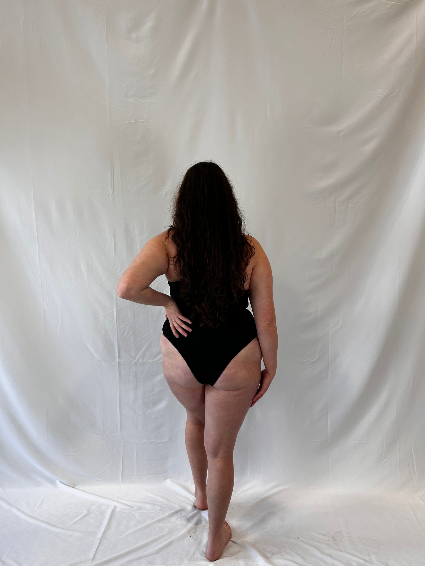 One piece in black from the back. Model wears the milkmaid style tie front crinkle fabric one piece. Black one piece is high cut and cheeky.