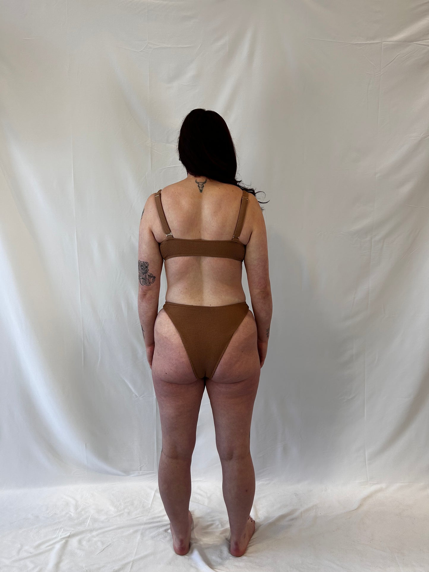 Model wears the milkmaid style tie front crinkle fabric bikini top. Model wears the high rise cheeky textured crinkle fabric bikini bottom in beige. Angle is from the back.
