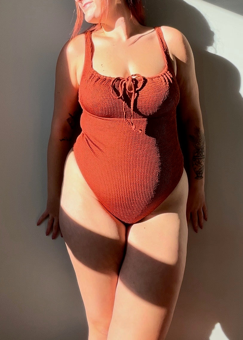 Close up of the rust one piece from the front. Swimsuit is the milkmaid style tie front crinkle fabric one piece. Rust one piece is high cut and cheeky.