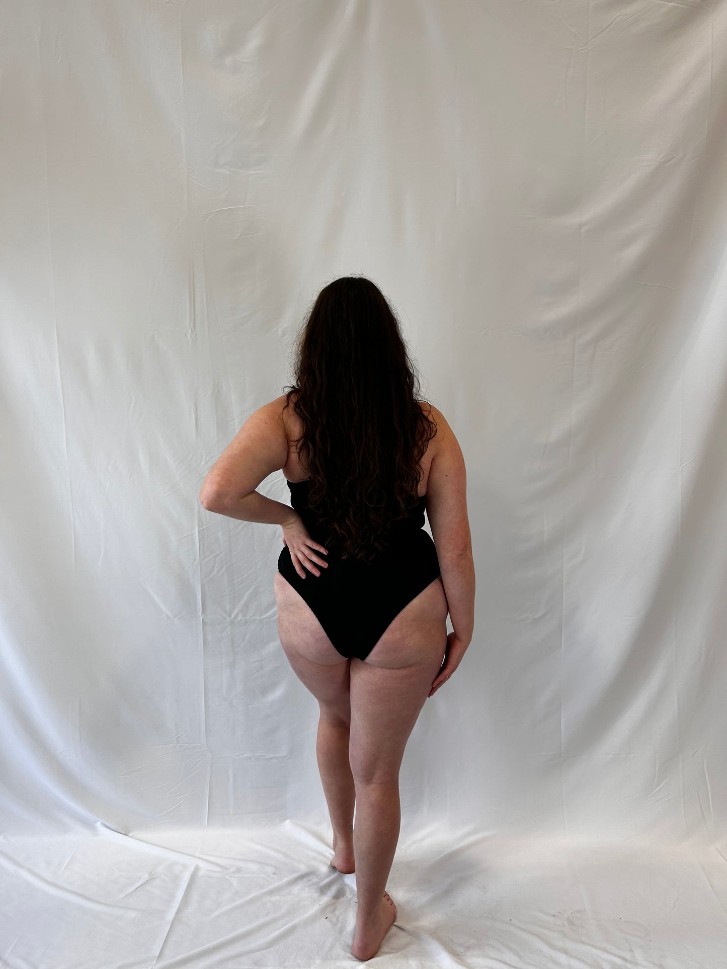 One piece in black from the back. Model wears the milkmaid style tie front crinkle fabric one piece. Black one piece is high cut and cheeky. 