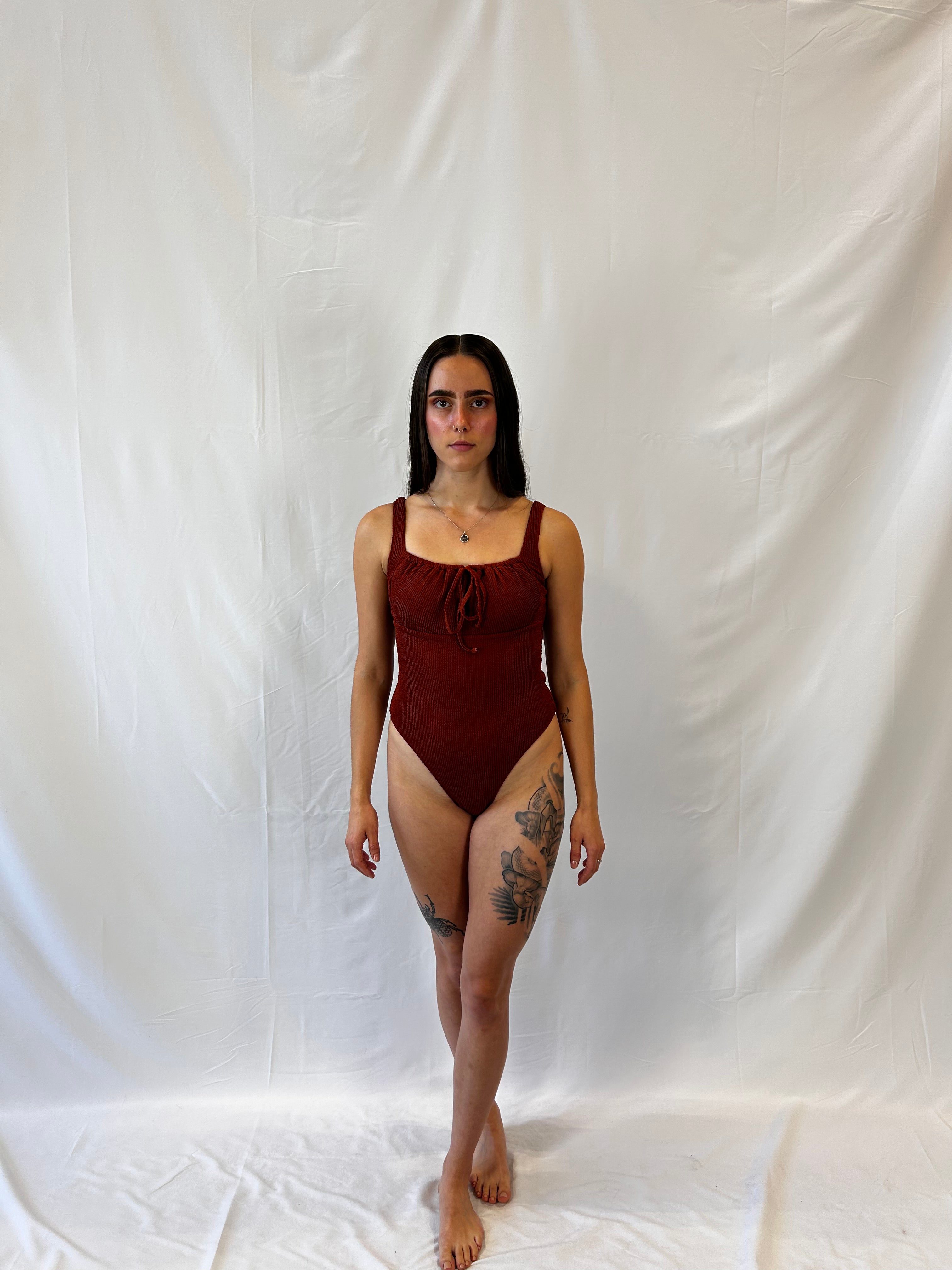 Front view of model wearing the one piece in rust. Woman wears the milkmaid style tie front crinkle fabric one piece. Rust one piece is high cut and cheeky.  