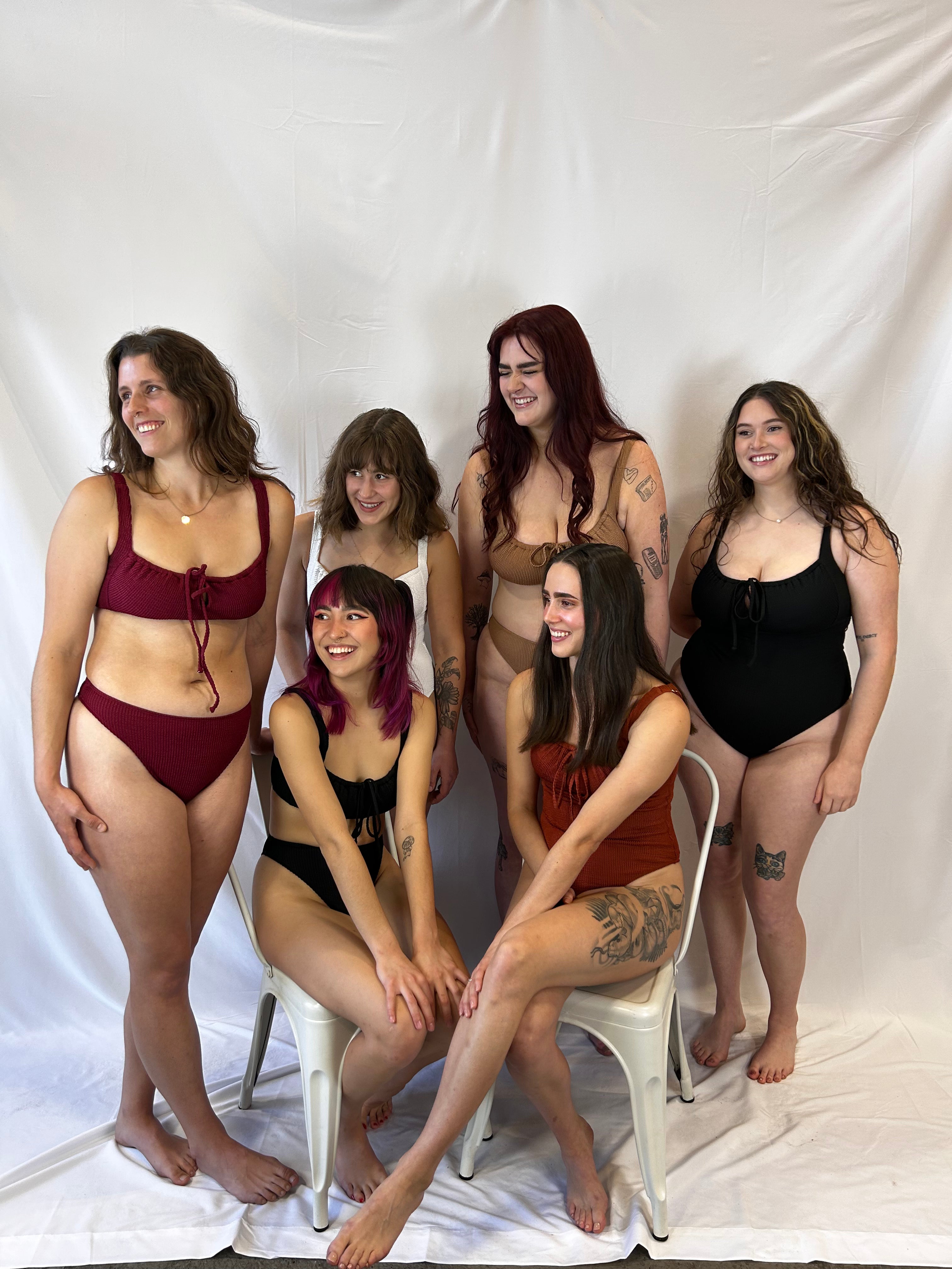 Models wearing (from left to right) the burgundy bikini, black bikini, white one piece, beige bikini, rust one piece, and black one piece swimsuits.