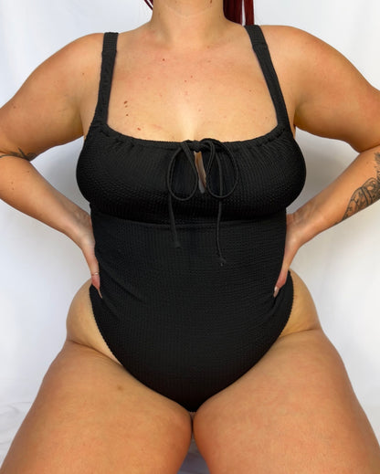 Close up of the black one piece from the front. Swimsuit is the milkmaid style tie front crinkle fabric one piece. Black one piece is high cut and cheeky. 