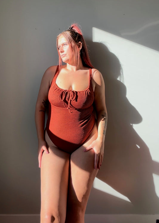 Close up view of model wearing the one piece in rust. Woman wears the milkmaid style tie front crinkle fabric one piece. Rust one piece is high cut and cheeky.